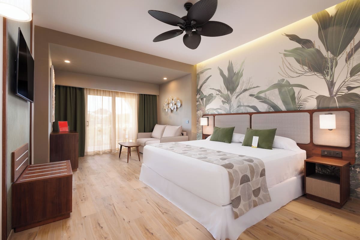 A hotel room with a large bed, a TV, a ceiling fan, a small table, and a seating area with a view of the outdoors.