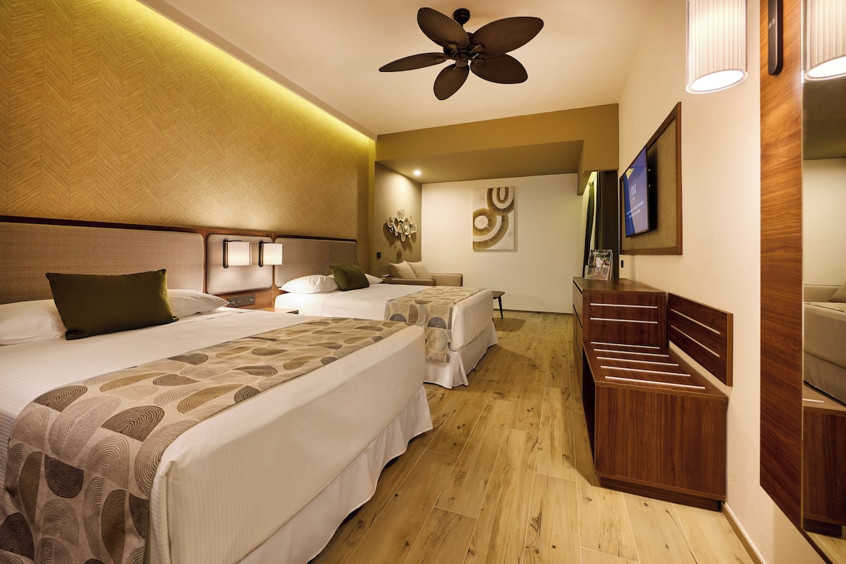 A hotel room with two beds, a wooden dresser, a ceiling fan, wall-mounted lamps, and a flat-screen TV.