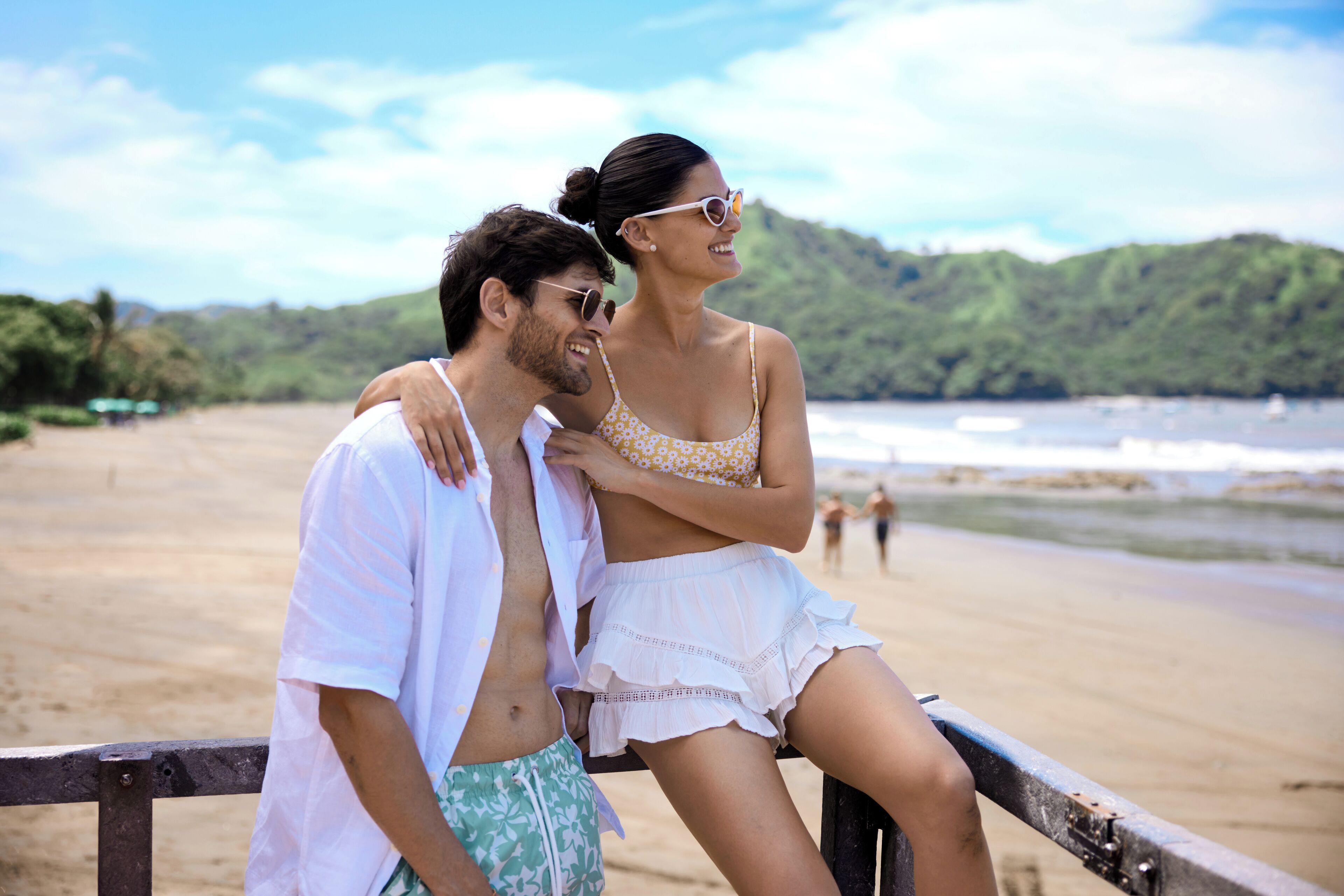 A couple sitting on a wooden bench overlooking a beach with a forested hill in the background.
