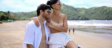 A couple sitting on a wooden bench overlooking a beach with a forested hill in the background.