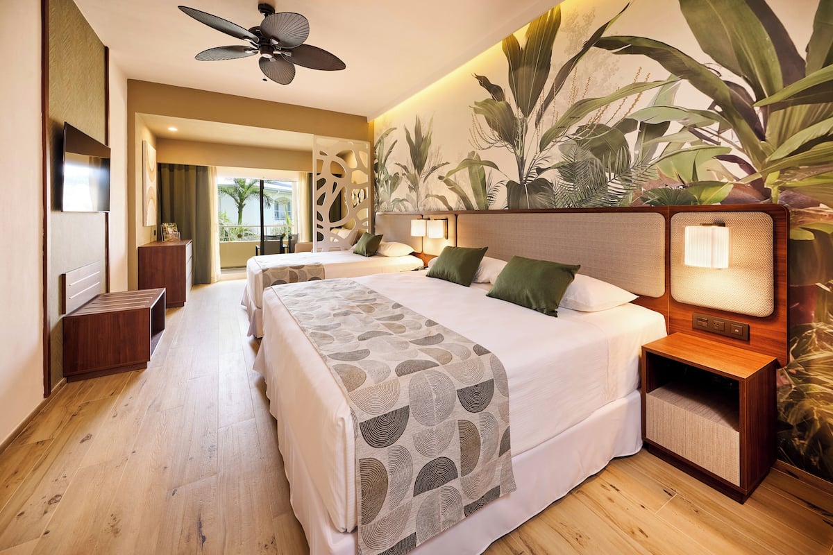 A hotel room with a large bed, two bedside tables, a ceiling fan, and a wall mural of tropical plants.