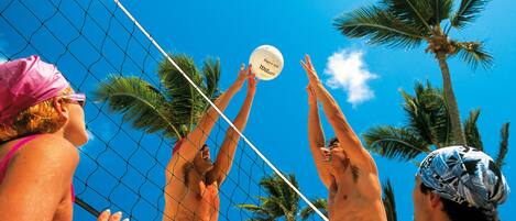 Un partido de voleibol de playa con jugadores que se estiran para alcanzar la pelota, palmeras y un cielo azul despejado.