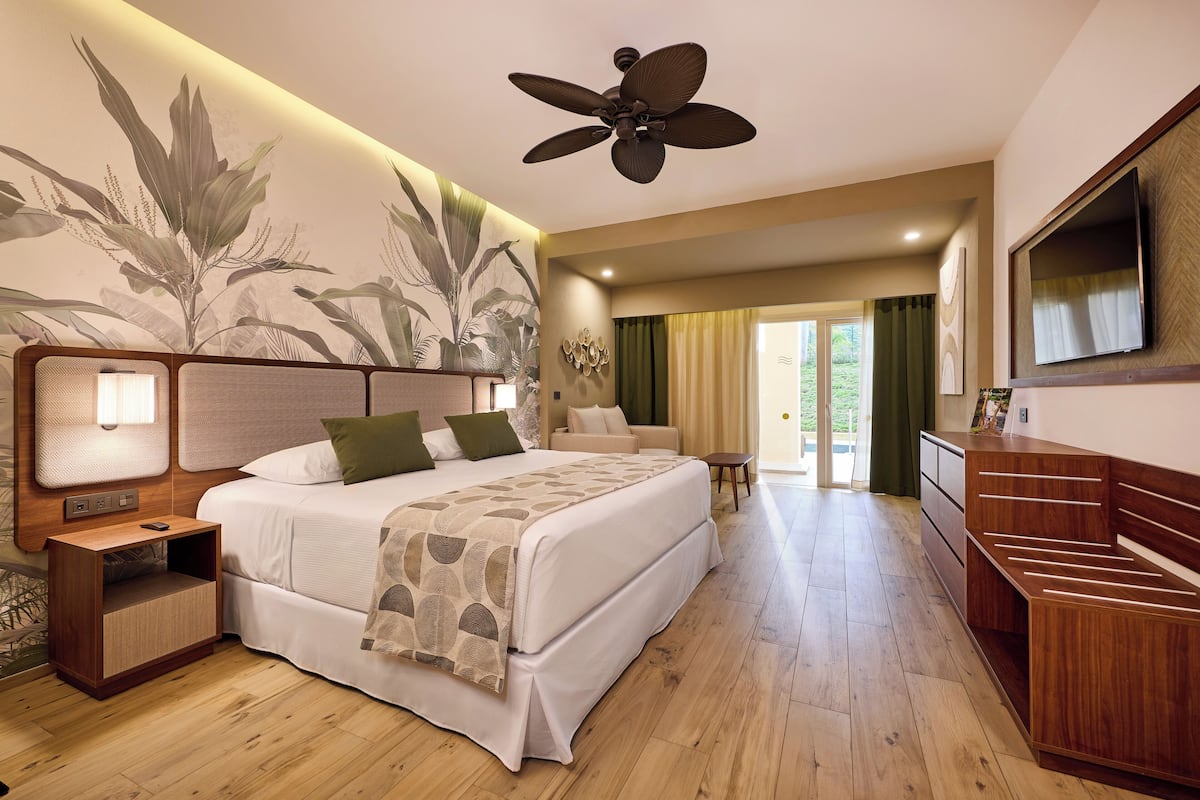 A spacious bedroom with a large bed, a nightstand, a wooden dresser, a ceiling fan, and a wall-mounted TV.