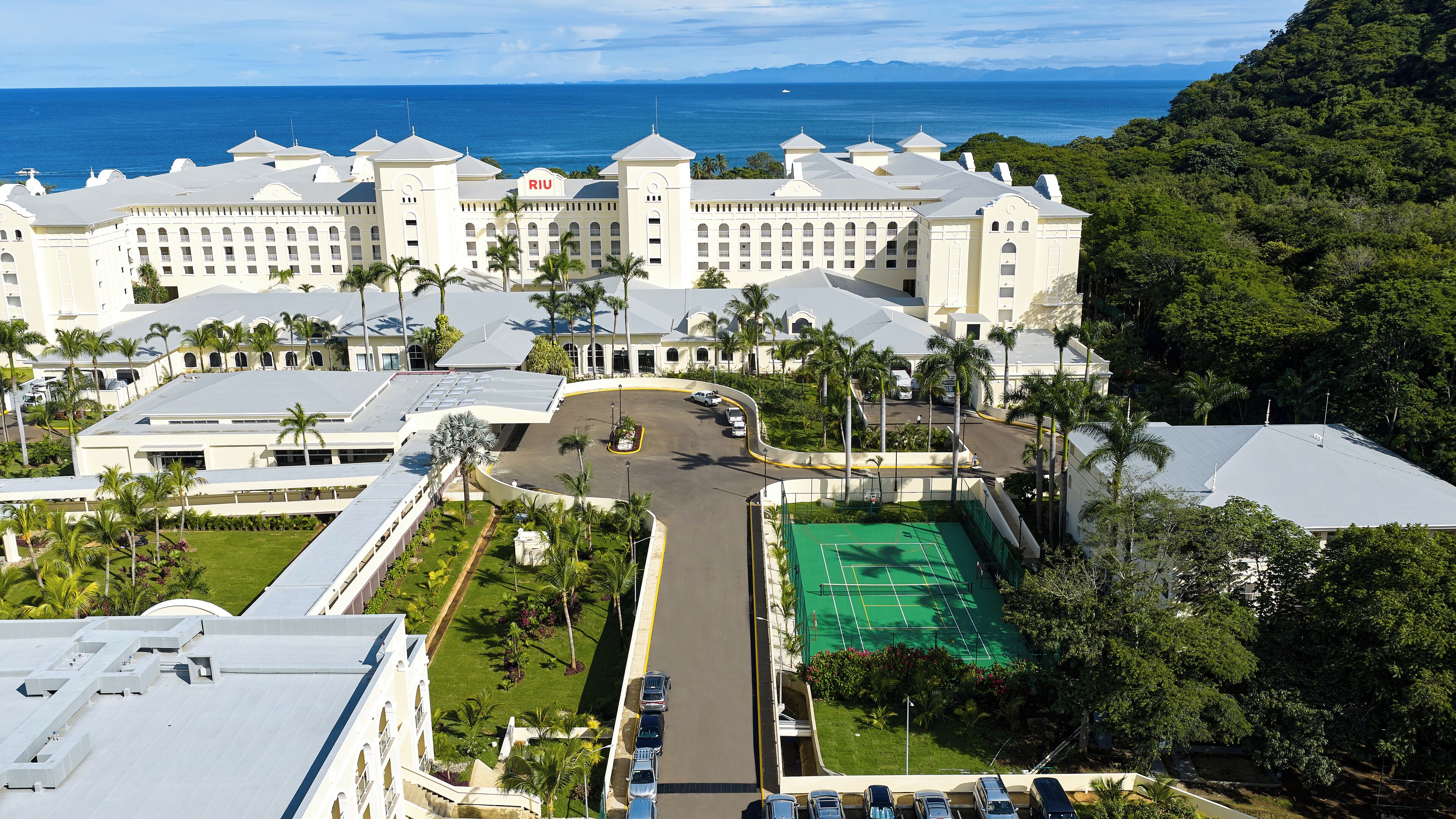 A large hotel with multiple wings, a tennis court, and a view of the ocean.