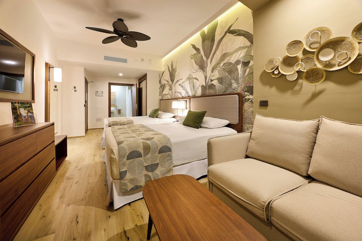 A hotel room with a bed, a sofa, a wooden table, a ceiling fan, and a wall with a circular pattern.
