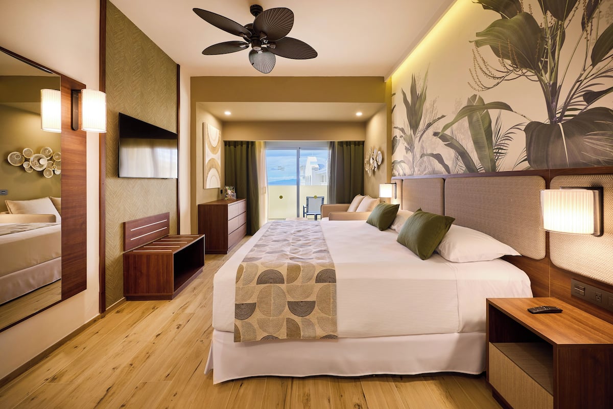 A hotel room with a large bed, a ceiling fan, a TV, and a balcony with a view.