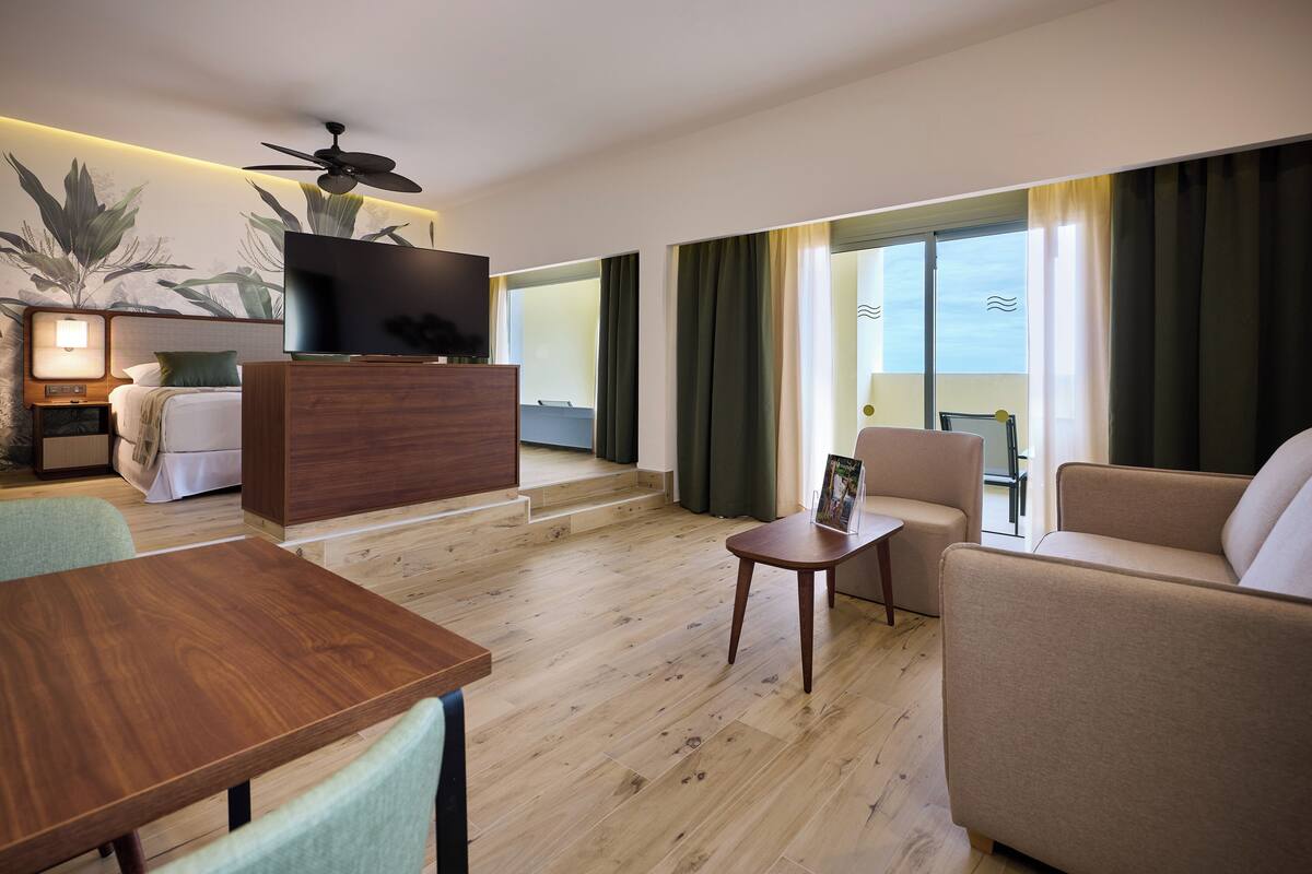 A modern hotel room with a wooden floor, a TV on a wooden cabinet, a sofa, a small table, and a balcony with curtains.