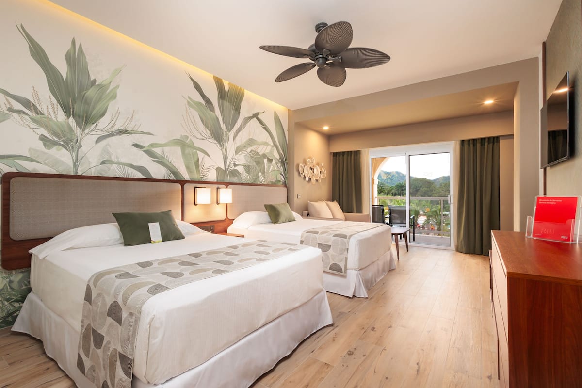 A hotel room with two beds, a ceiling fan, a TV, and a balcony with a view of the mountains.