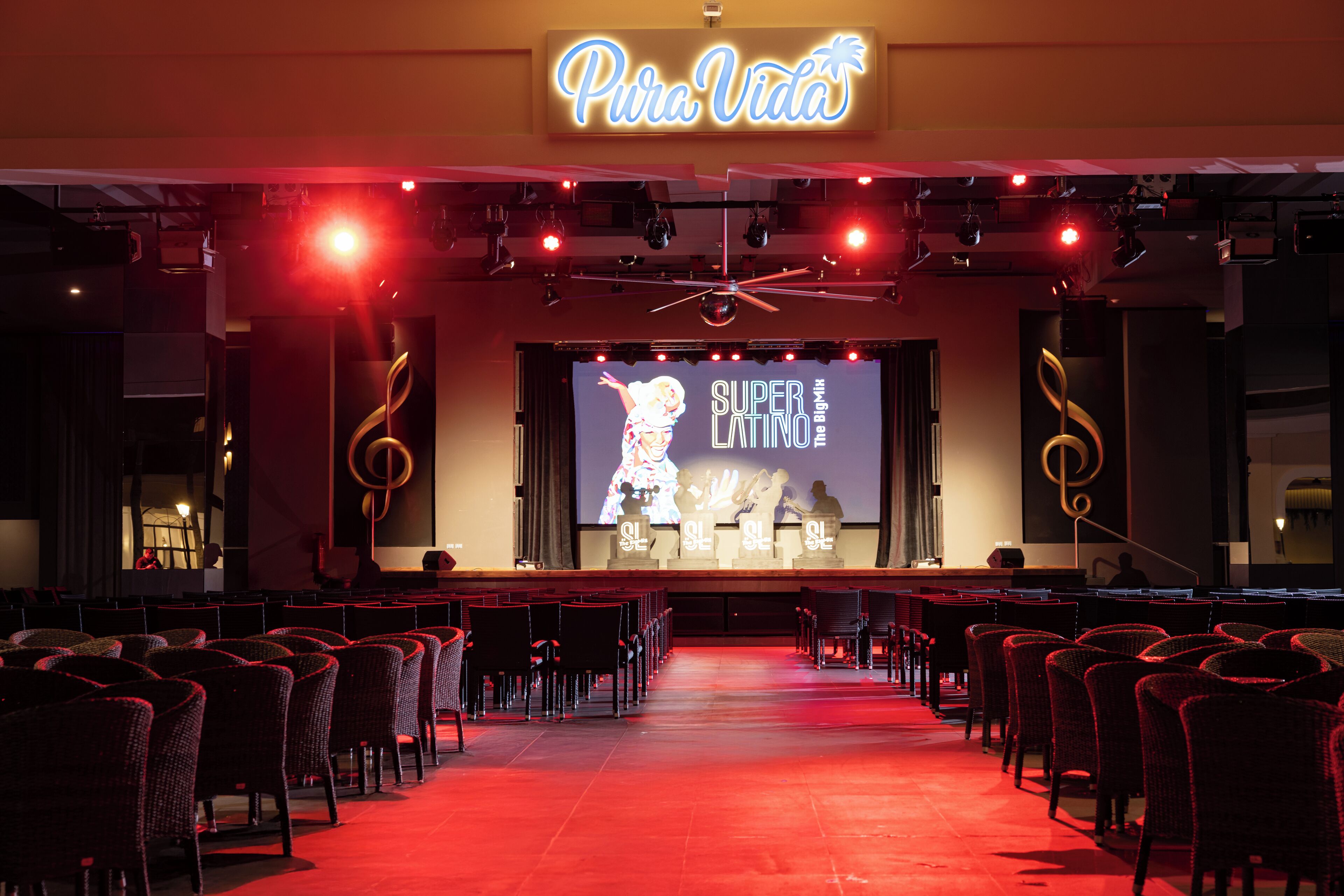 An auditorium with rows of chairs facing a stage, a large screen displaying 'Super Latino The Big Show', and a 'Pura Vida' sign above.
