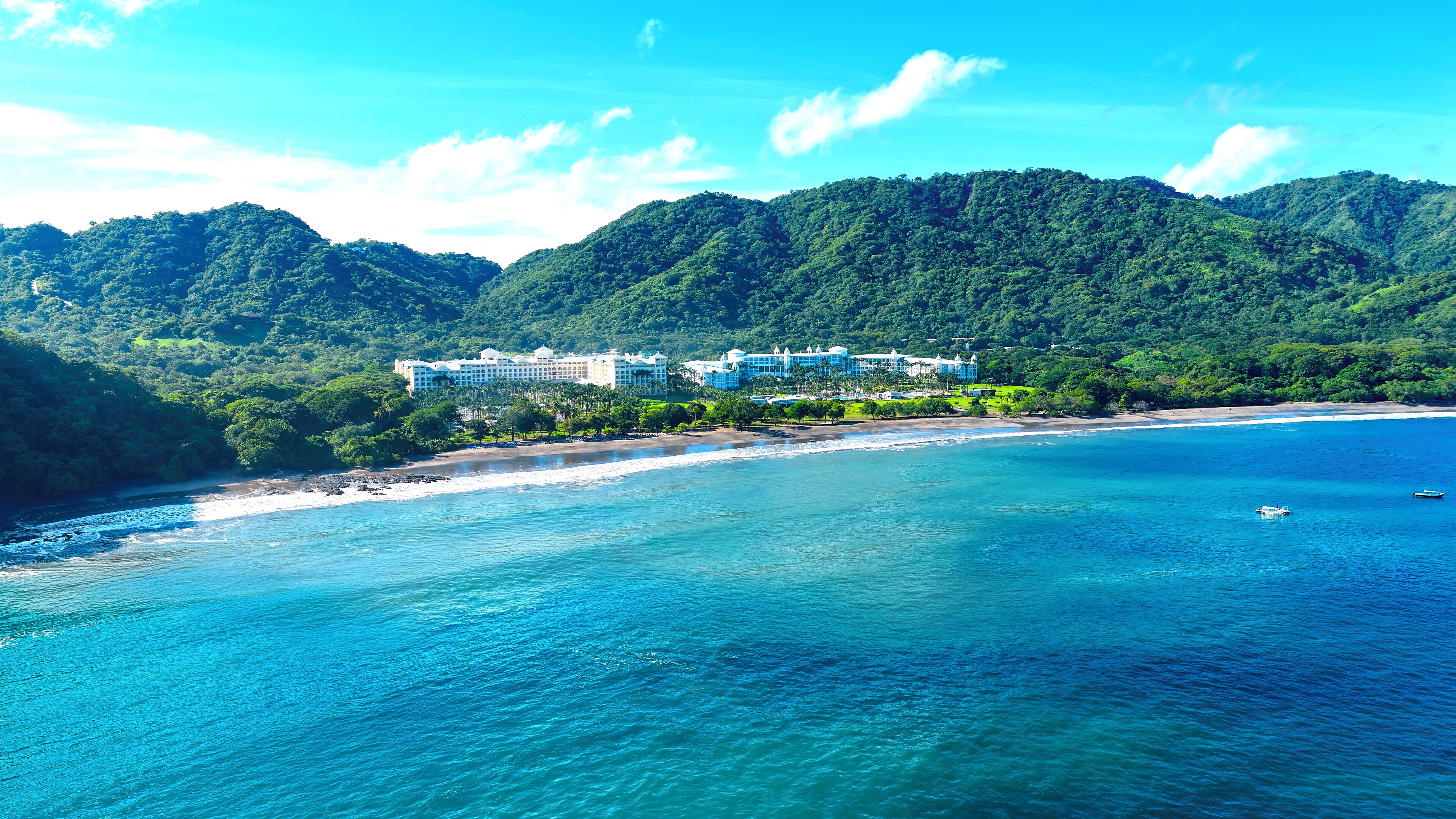 A coastal resort with multiple buildings, a beach, and lush green mountains in the background.