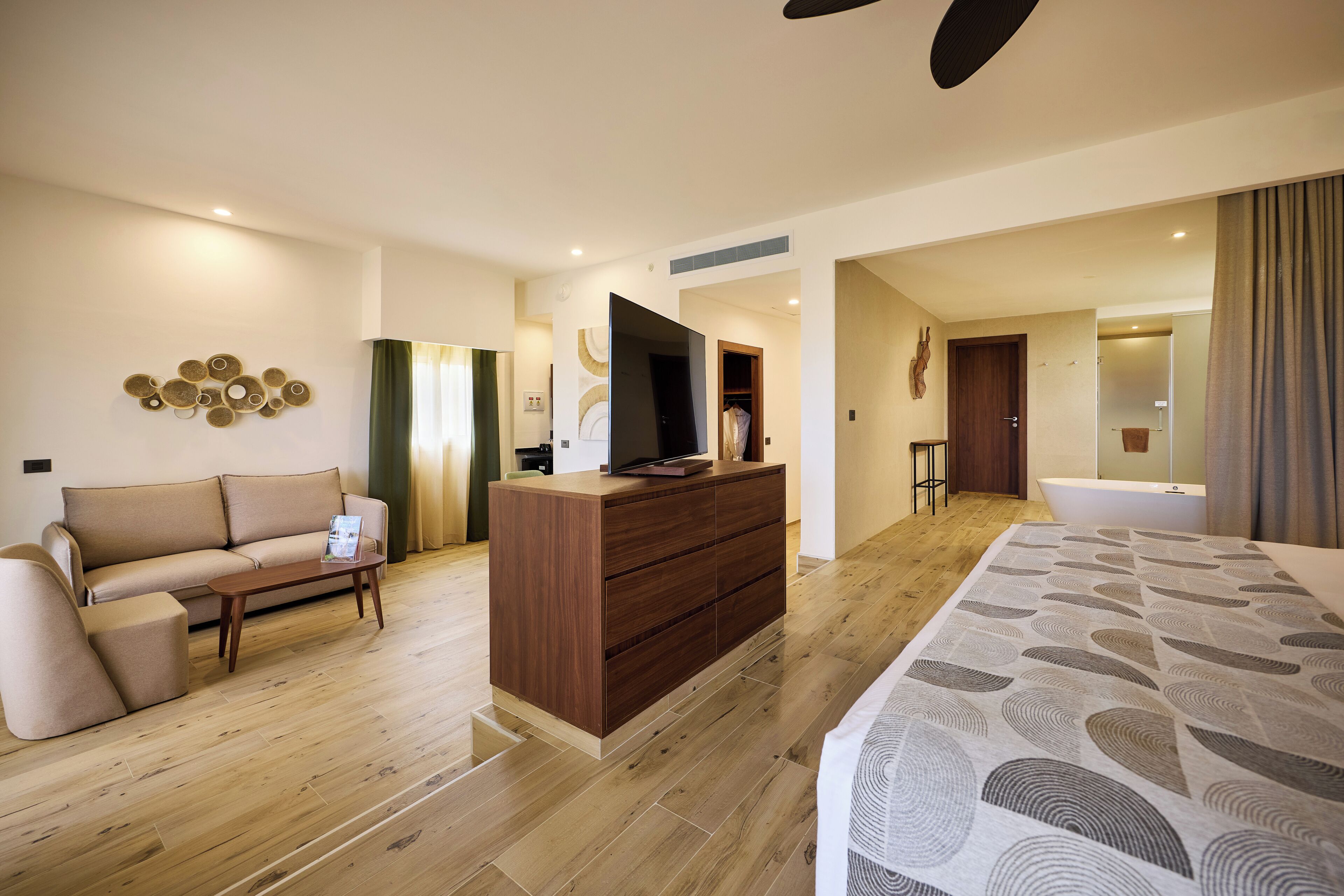 A modern hotel room with a wooden floor, a sofa, a TV on a wooden cabinet, and a large bed.
