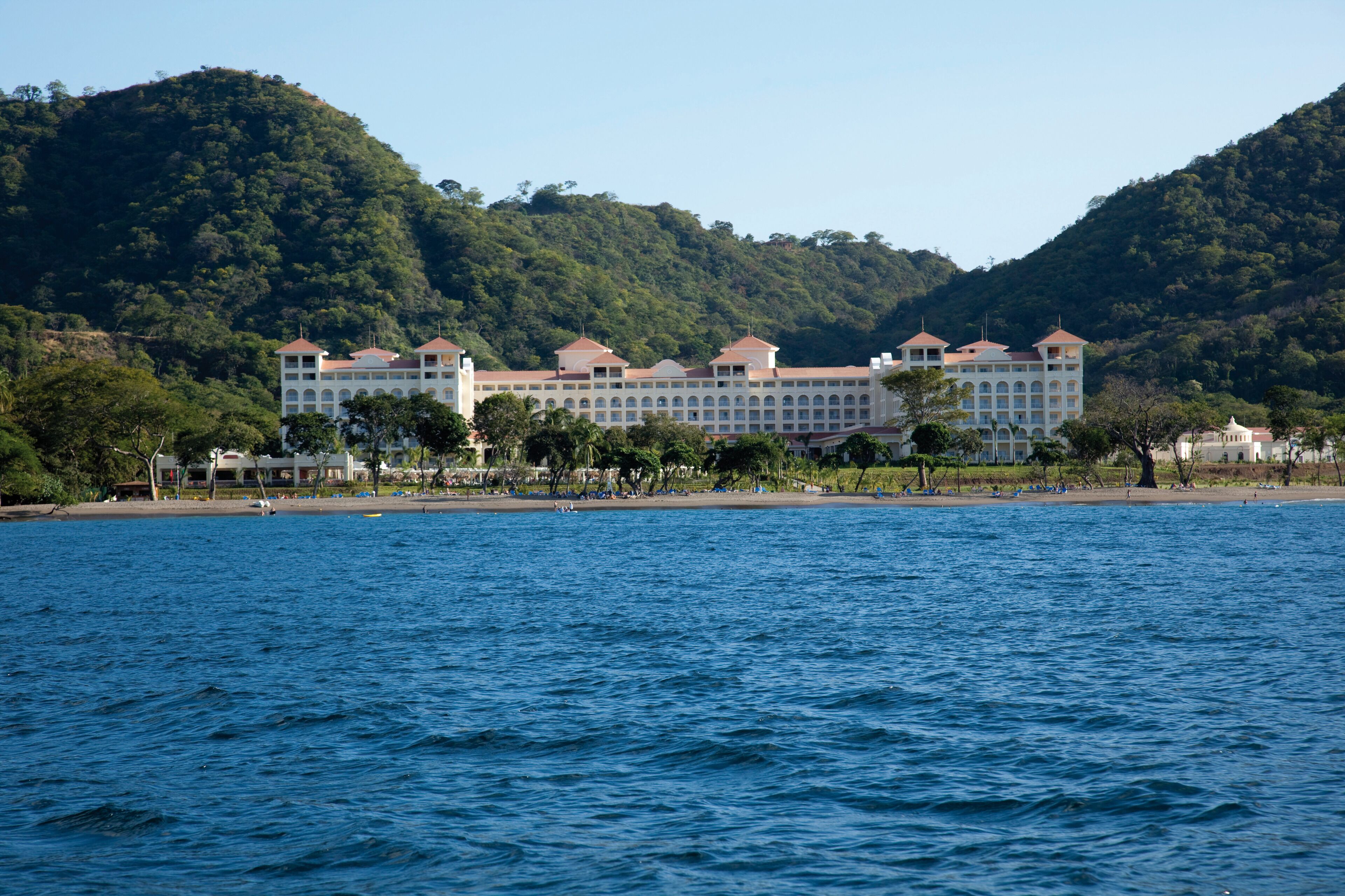 Un gran hotel con varias edificaciones, una playa en primer plano y colinas al fondo.