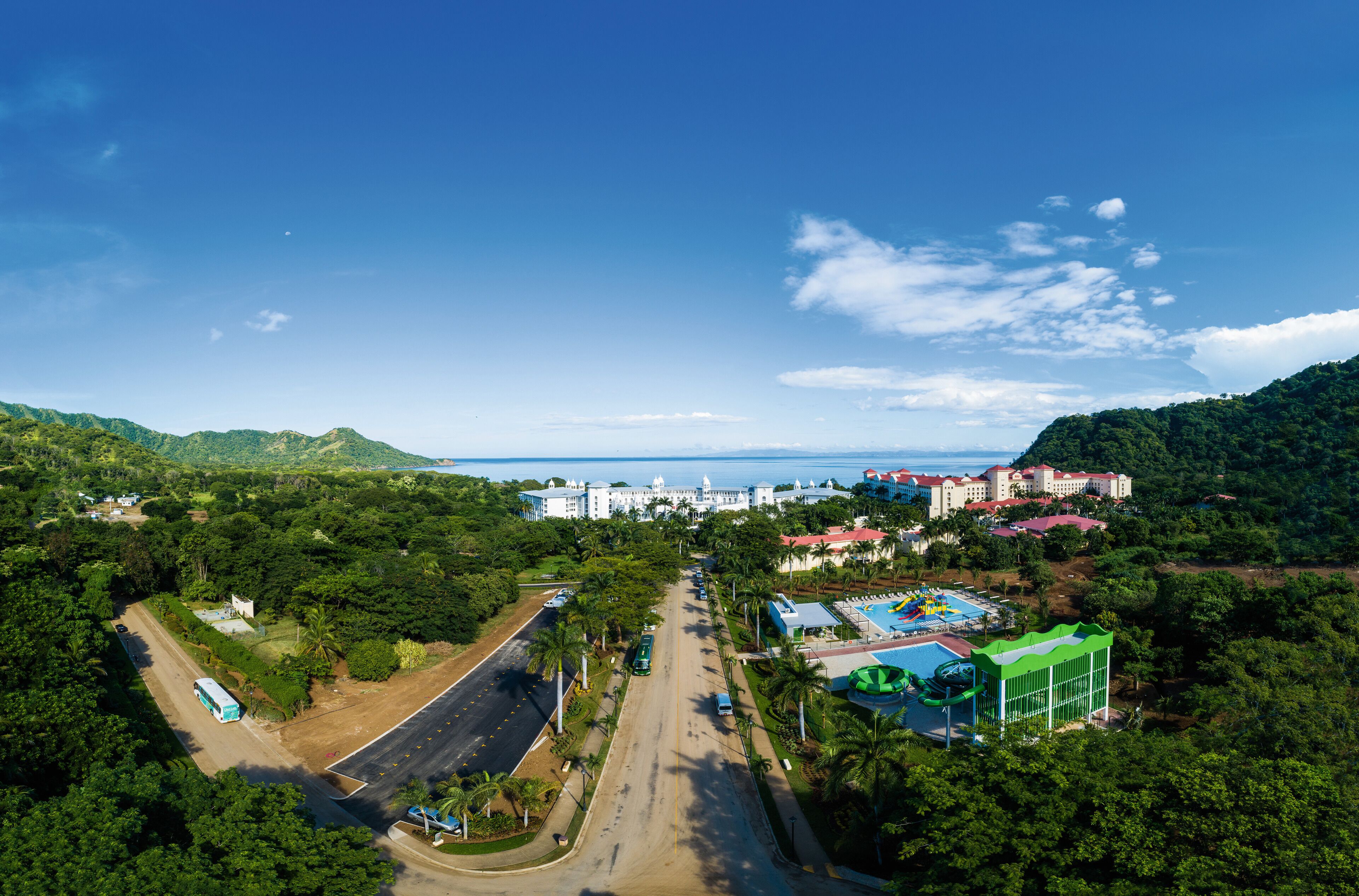 A resort with a swimming pool, a large building, and a road leading to the beach.
