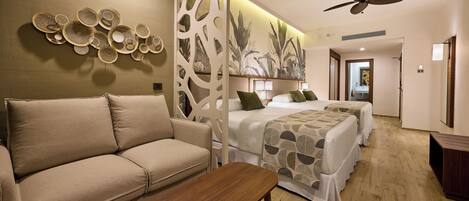 A hotel room with a beige sofa, a wooden bench, two beds with patterned bedding, a decorative wall with circular designs, a ceiling fan, and a wooden floor.