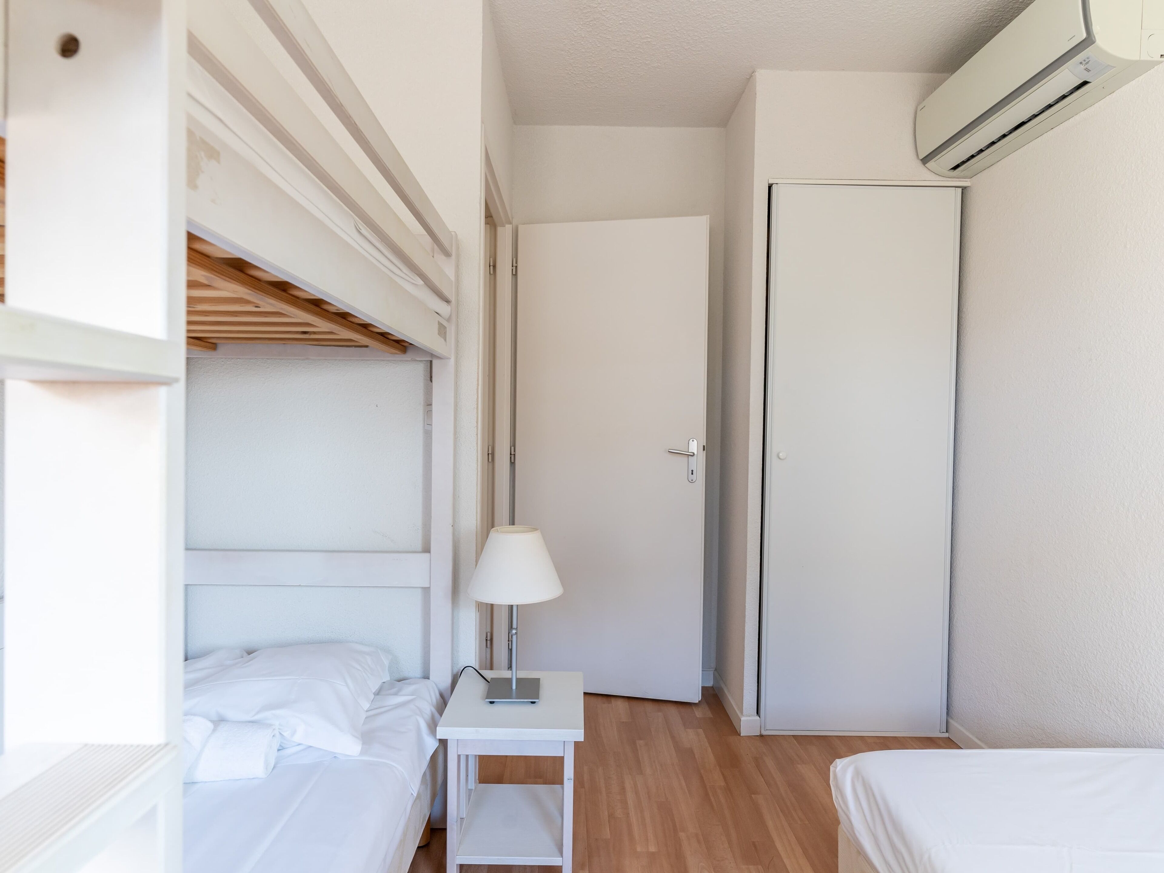 A compact hotel room with a bunk bed, a bedside table with a lamp, a wardrobe, and an air conditioning unit.