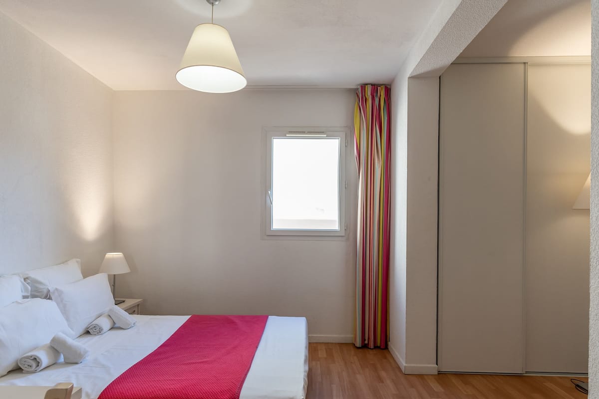 A hotel room with a bed, a window, a wardrobe, and a bedside table with a lamp.