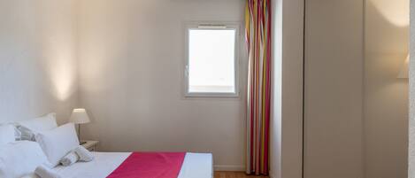 A hotel room with a bed, a window, a wardrobe, and a bedside table with a lamp.