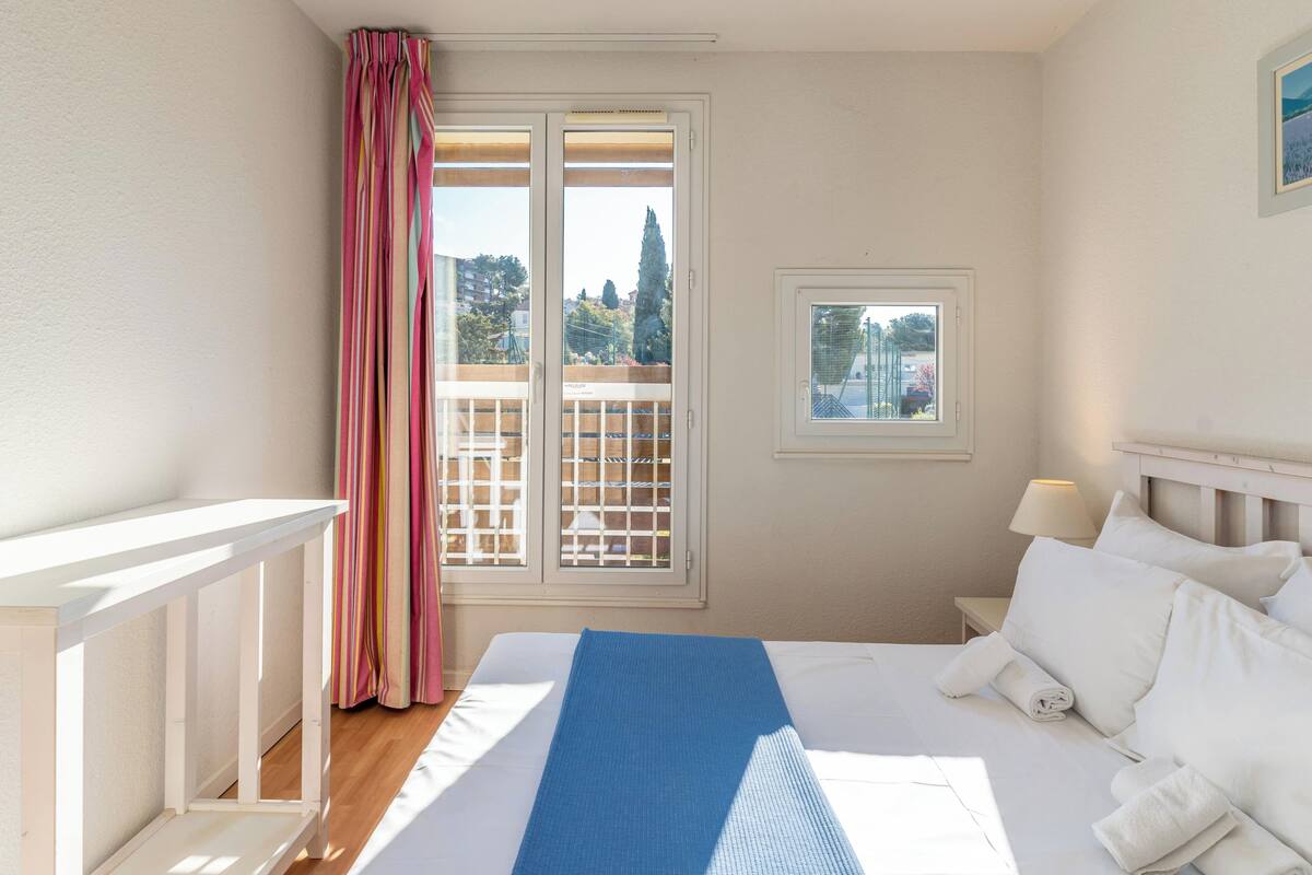A hotel room with a bed, a small window, a balcony view, and a curtain.