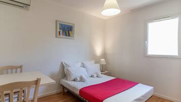Una cama individual con ropa de cama blanca y una manta roja, una mesita de noche con lámpara, una silla de madera, un cuadro en la pared y una ventana con luz natural.
