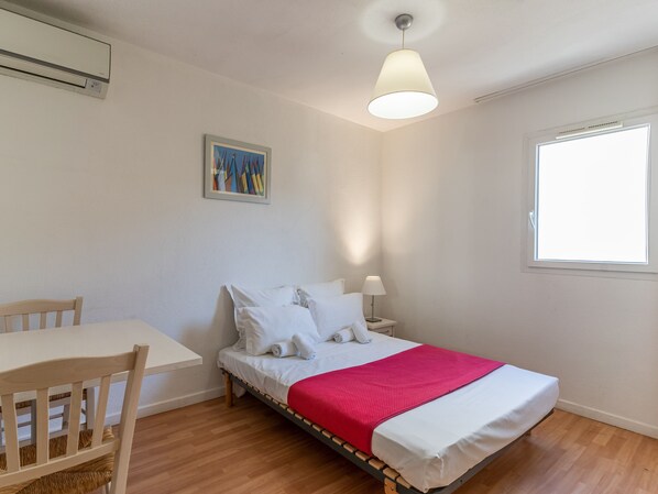 Una cama individual con ropa de cama blanca y una manta roja, una mesita de noche con lámpara, una silla de madera, un cuadro en la pared y una ventana con luz natural.