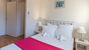 A neatly made bed with white bedding and a pink blanket, two bedside lamps, a small bedside table, and a painting on the wall. - Vacanceole Residence L'Ile d'Or (La Londe-les-Maures)