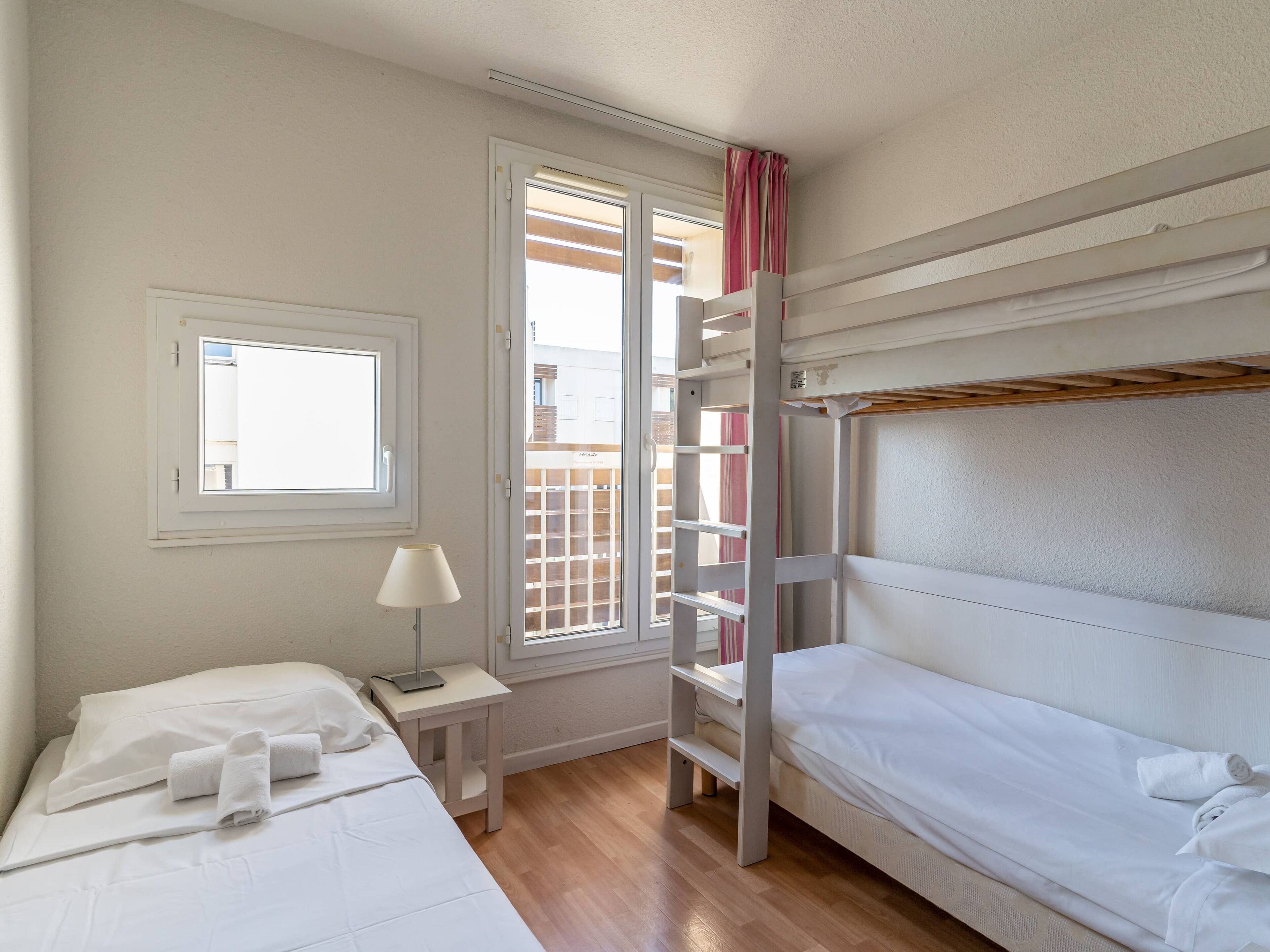 Una habitación pequeña y limpia con una cama nido, una mesita de noche con lámpara y una ventana con persianas.