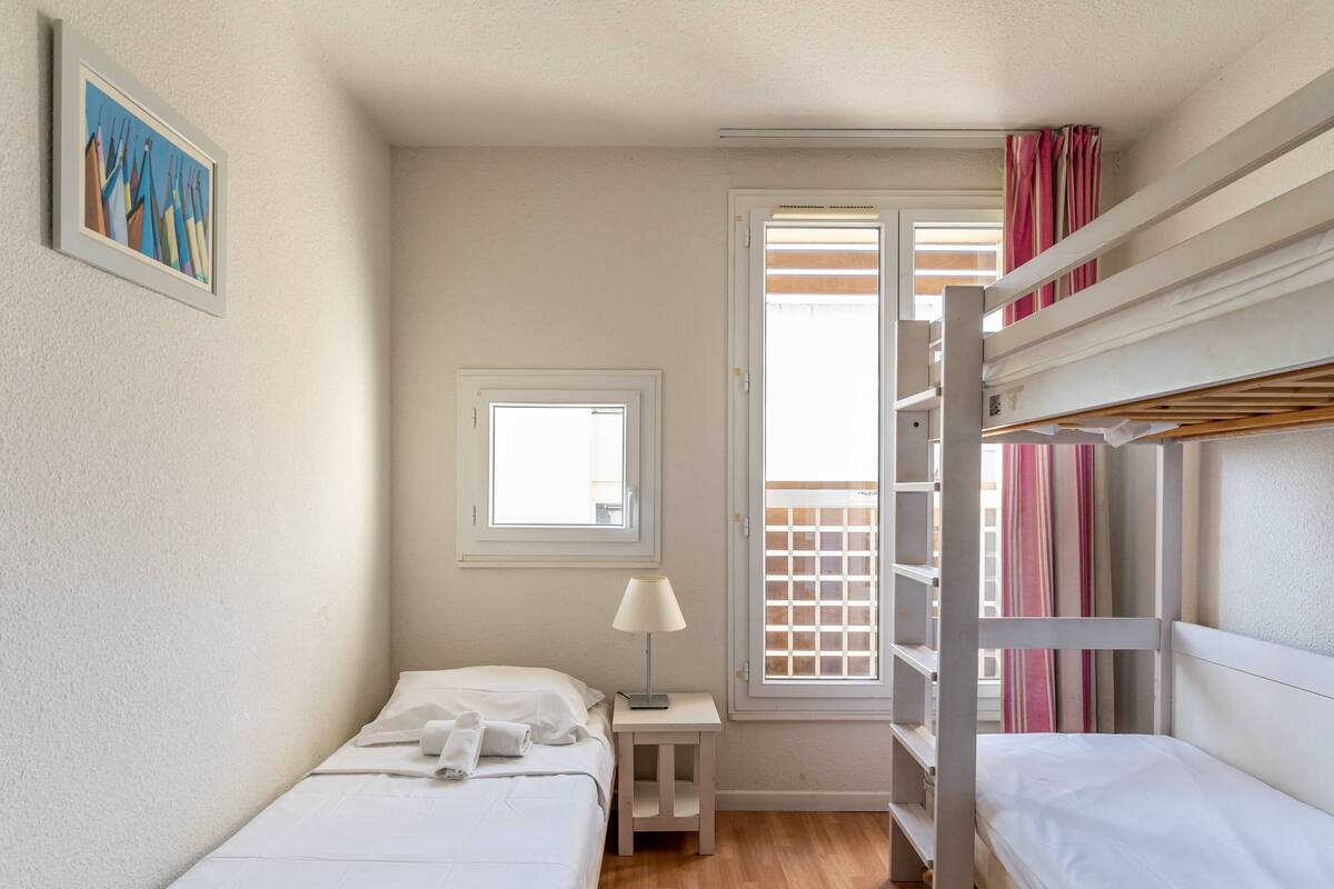 A small, well-lit room with a bunk bed, a single bed, a nightstand, a lamp, and a window.