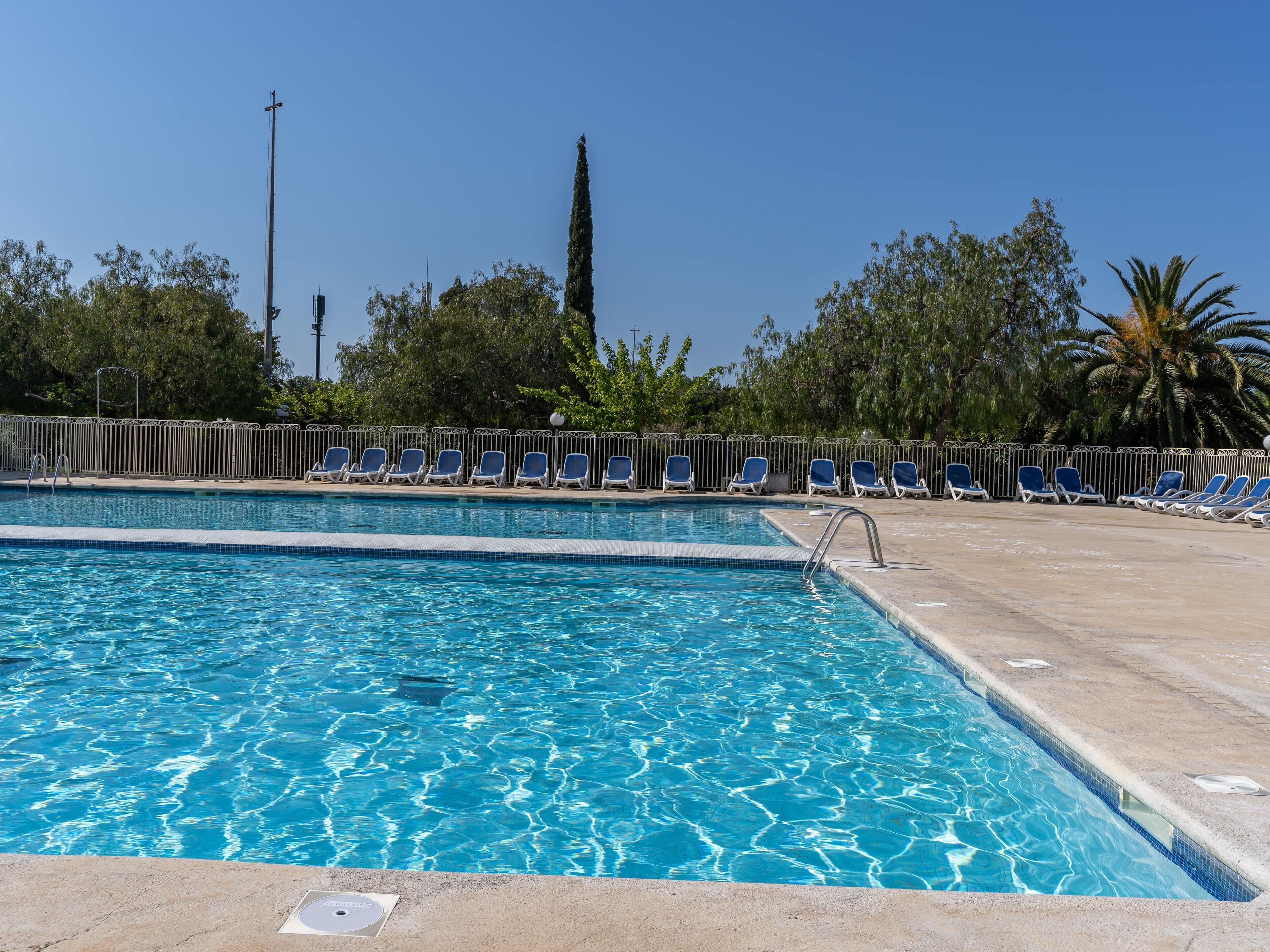 A swimming pool with clear blue water, surrounded by a metal fence and blue lounge chairs.