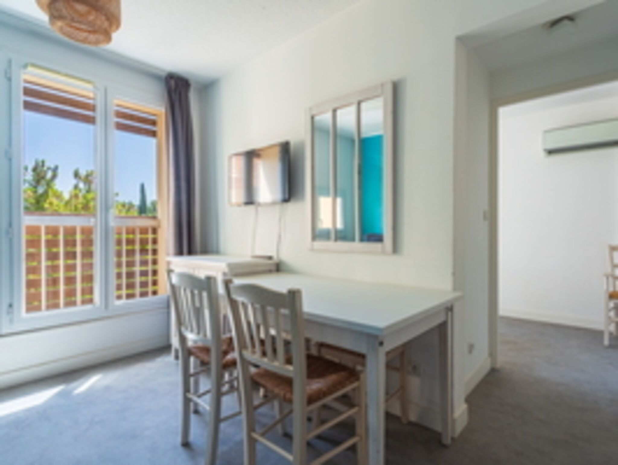 Una habitación con una mesa blanca, dos sillas, un televisor montado en la pared y una ventana con vistas a la vegetación.