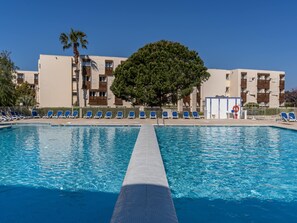 A swimming pool with blue water, surrounded by lounge chairs and a building with multiple balconies.