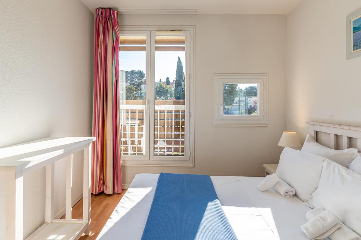 A hotel room with a bed, a small window, a balcony view, and a curtain.