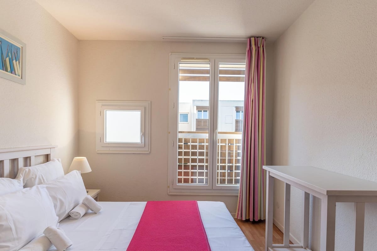 A hotel room with a bed, a window, a door, and a view of buildings outside.