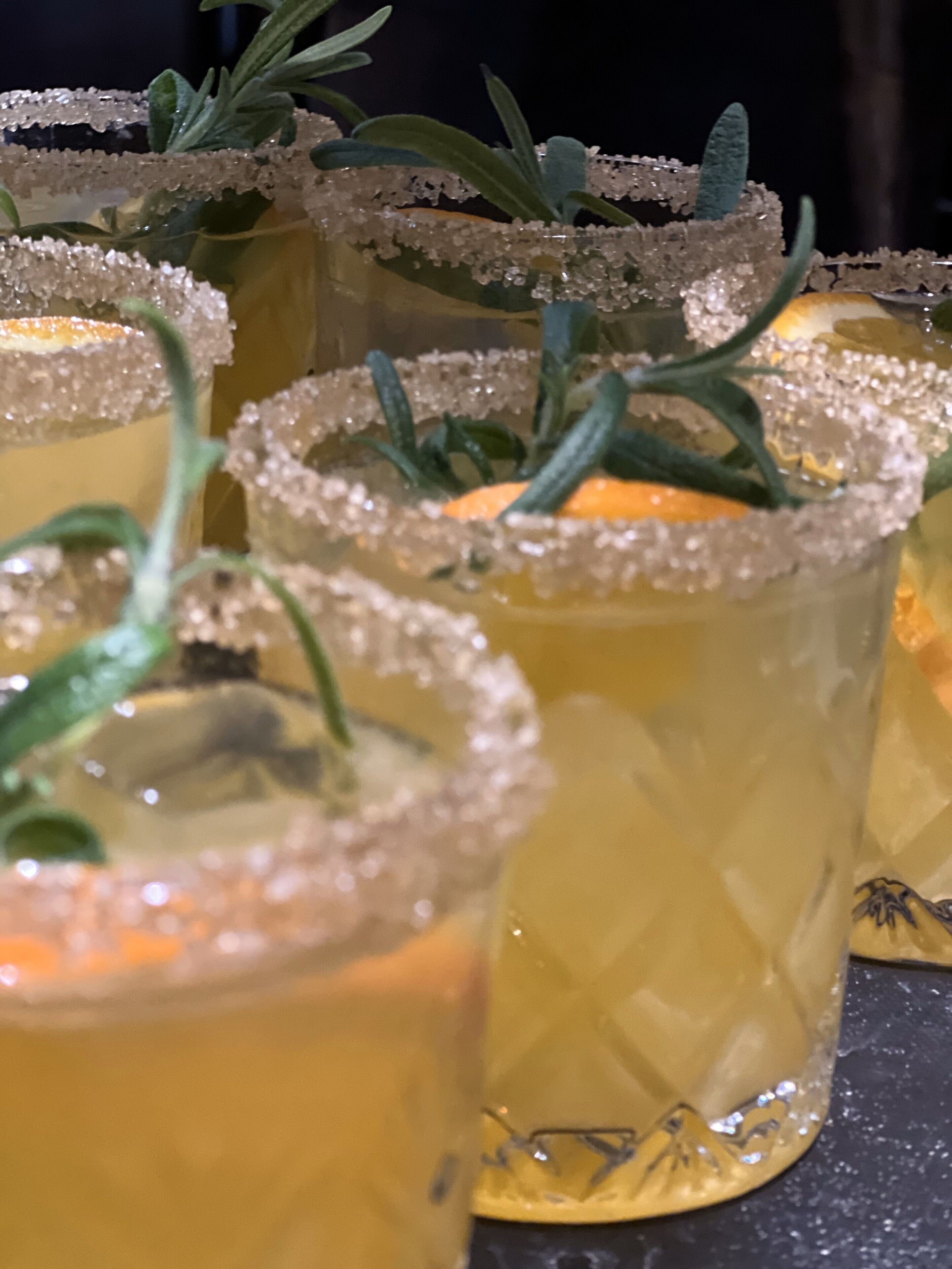 A close-up of several glasses of a yellow beverage garnished with rosemary and sugar.