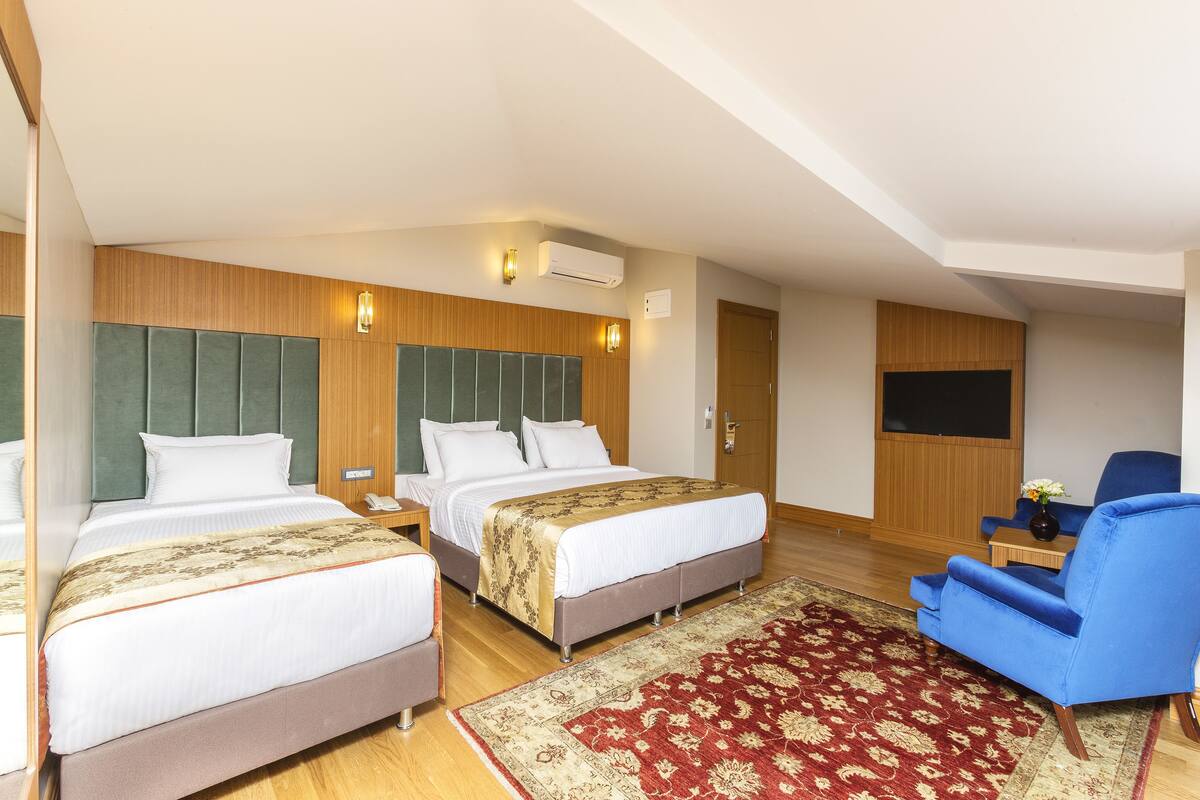 Habitación de hotel con dos camas, un sillón azul, cabecero de madera, alfombra roja estampada y televisor de pantalla plana.