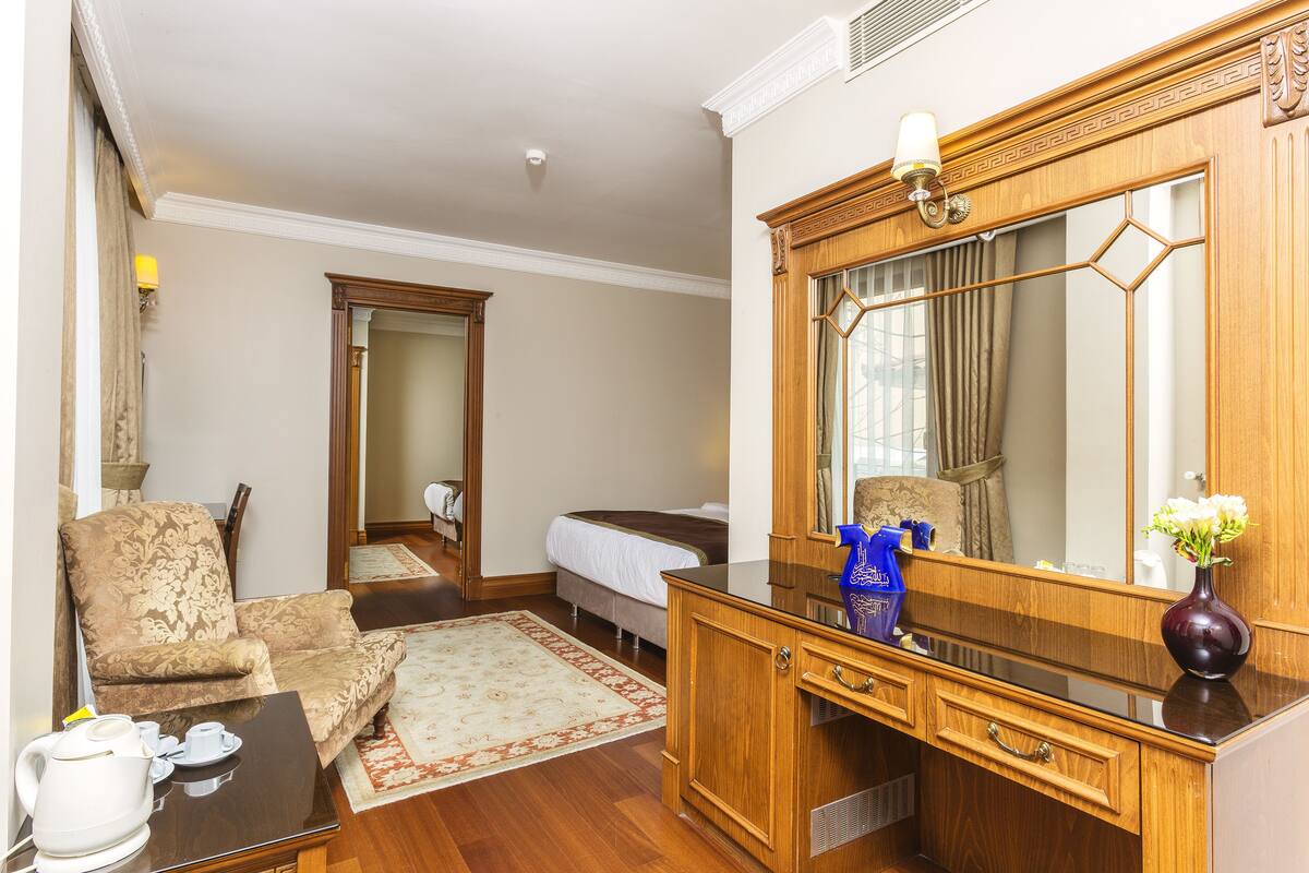 Habitación de hotel con cama, zona de estar con una silla y una mesa, cómoda de madera, espejo grande y un jarrón con flores.
