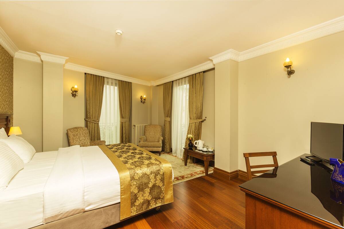 Habitación de hotel con cama, dos sillones, escritorio con televisor y una mesita de noche con lámpara y florero.