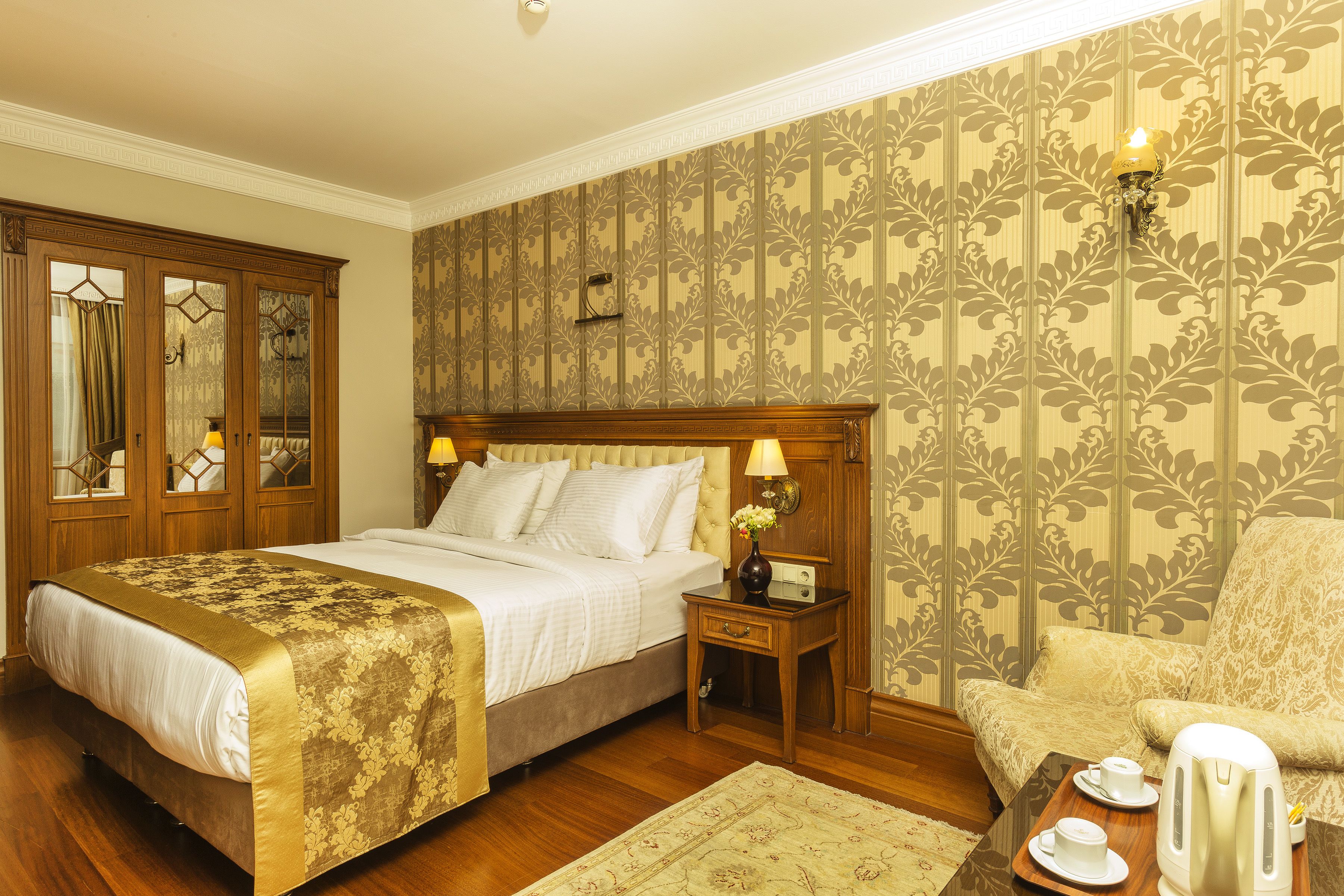 A bedroom with a large bed, a nightstand with a lamp, a chair, and a tray with a kettle and cups.