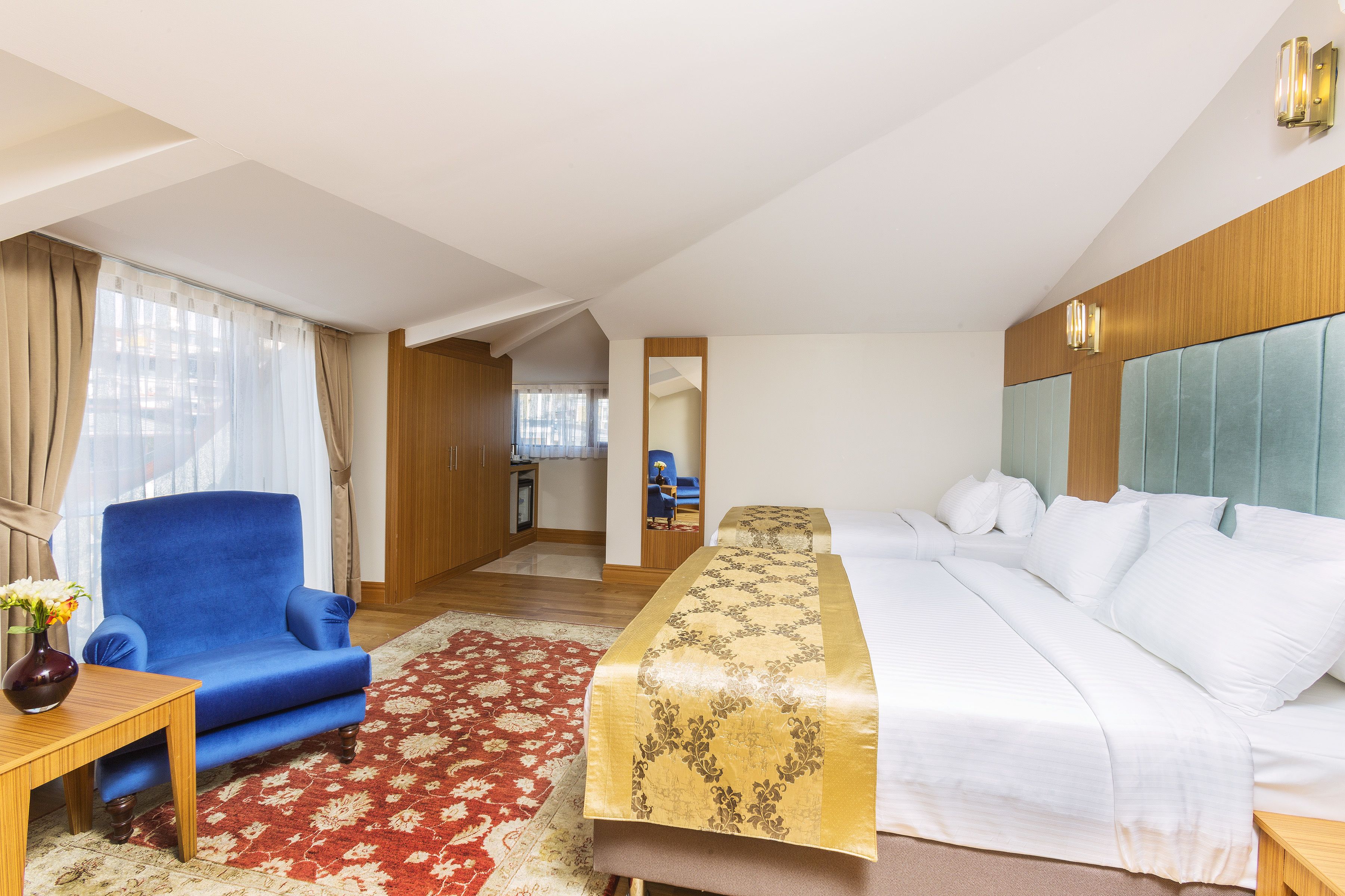 A hotel room with a bed, a blue armchair, a wooden side table, a red and white patterned rug, and a view of the hallway.
