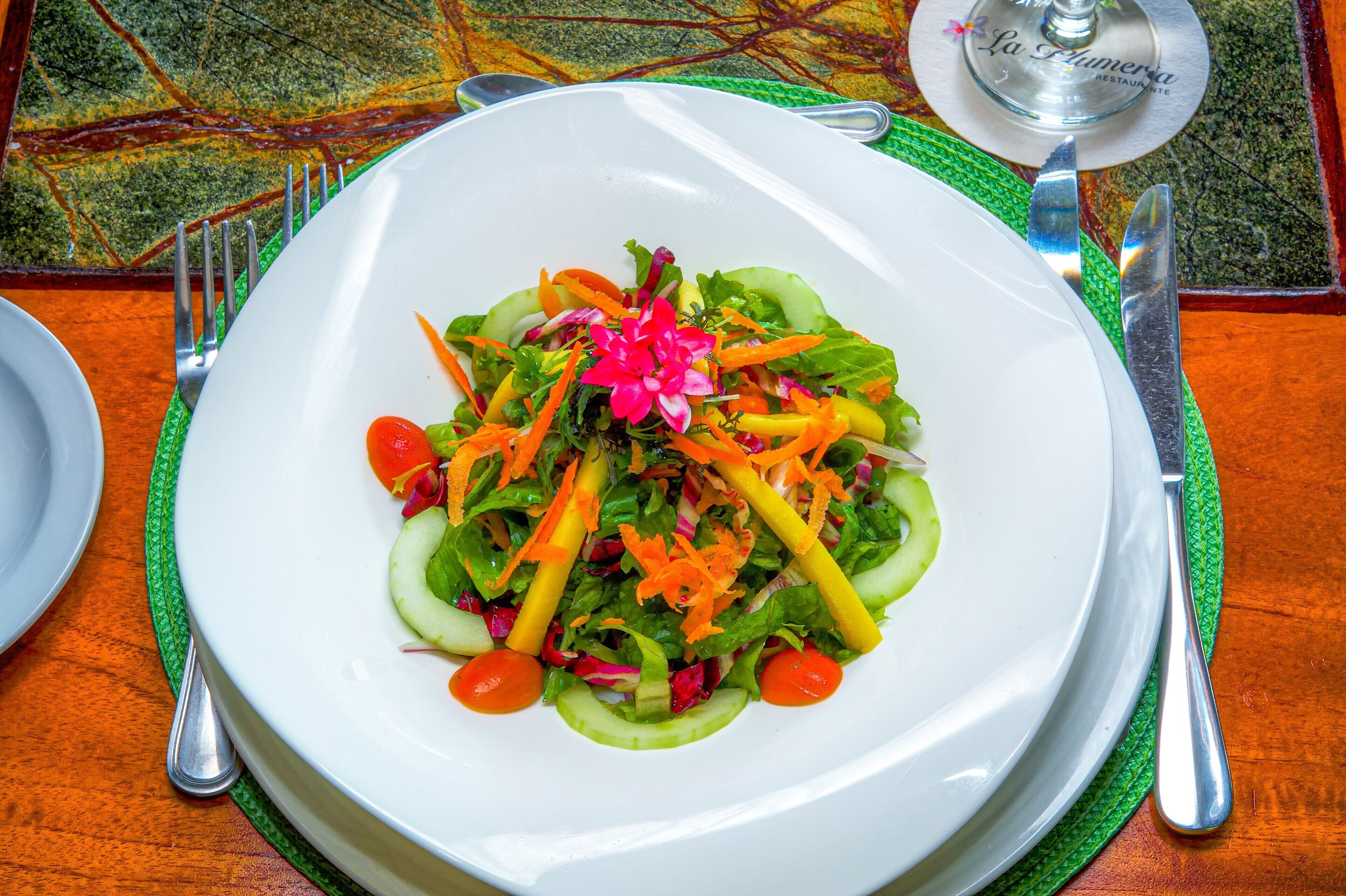 Ein weißer Teller mit einem farbenfrohen Salat auf einer grünen Tischmatte, dazu Besteck und ein Glas auf einem Tisch mit gemustertem Tuch.