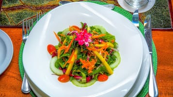 Ein weißer Teller mit einem farbenfrohen Salat auf einer grünen Tischmatte, dazu Besteck und ein Glas auf einem Tisch mit gemustertem Tuch.