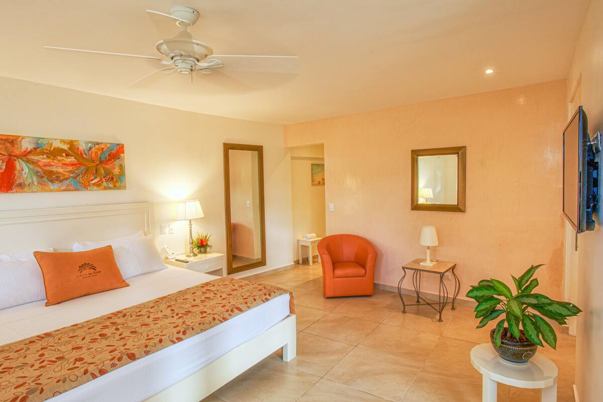 A hotel room with a bed, a ceiling fan, a chair, a small table with a lamp, and a potted plant.