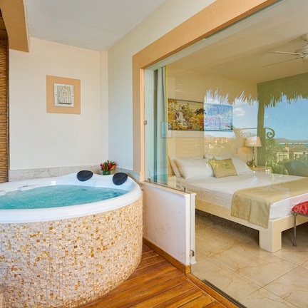 A room with a glass-enclosed balcony, a bed, a red bench, and a hot tub with mosaic tiles.