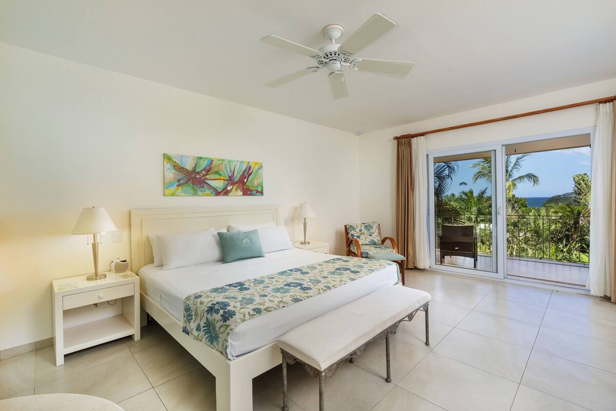 A spacious bedroom with a large bed, a ceiling fan, a nightstand, a bench, a chair, and a view of the outdoors.