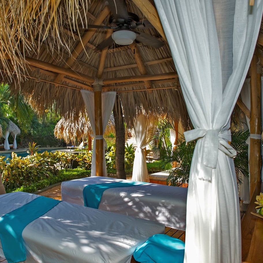A thatched-roof gazebo with two beds, each with a white bedspread and teal accents, surrounded by lush greenery.