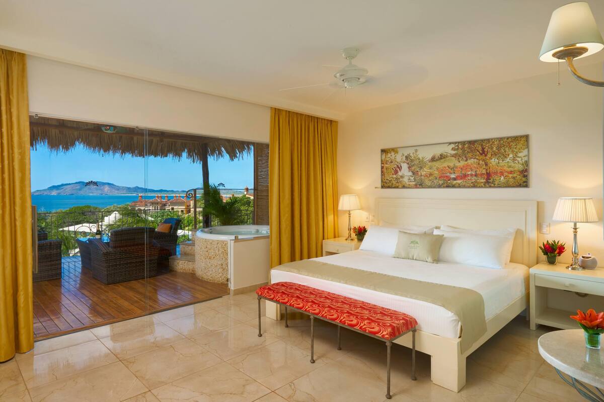 A hotel room with a large bed, a view of the ocean, a balcony with wicker furniture, and a painting on the wall.