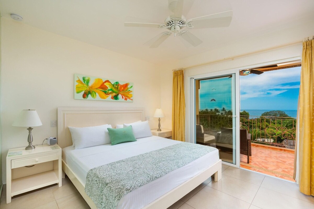 A hotel room with a bed, nightstands, a ceiling fan, a painting, and a balcony with a view.