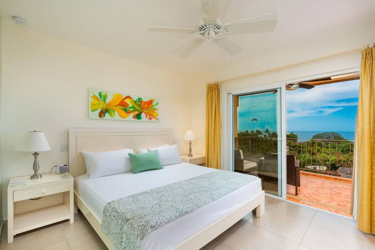 A hotel room with a bed, nightstands, a ceiling fan, a painting, and a balcony with a view.
