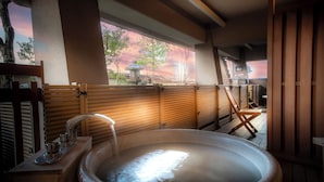 Un jacuzzi con un banco de madera y un farol de piedra, frente a un muro de madera y una vista de árboles y un atardecer.