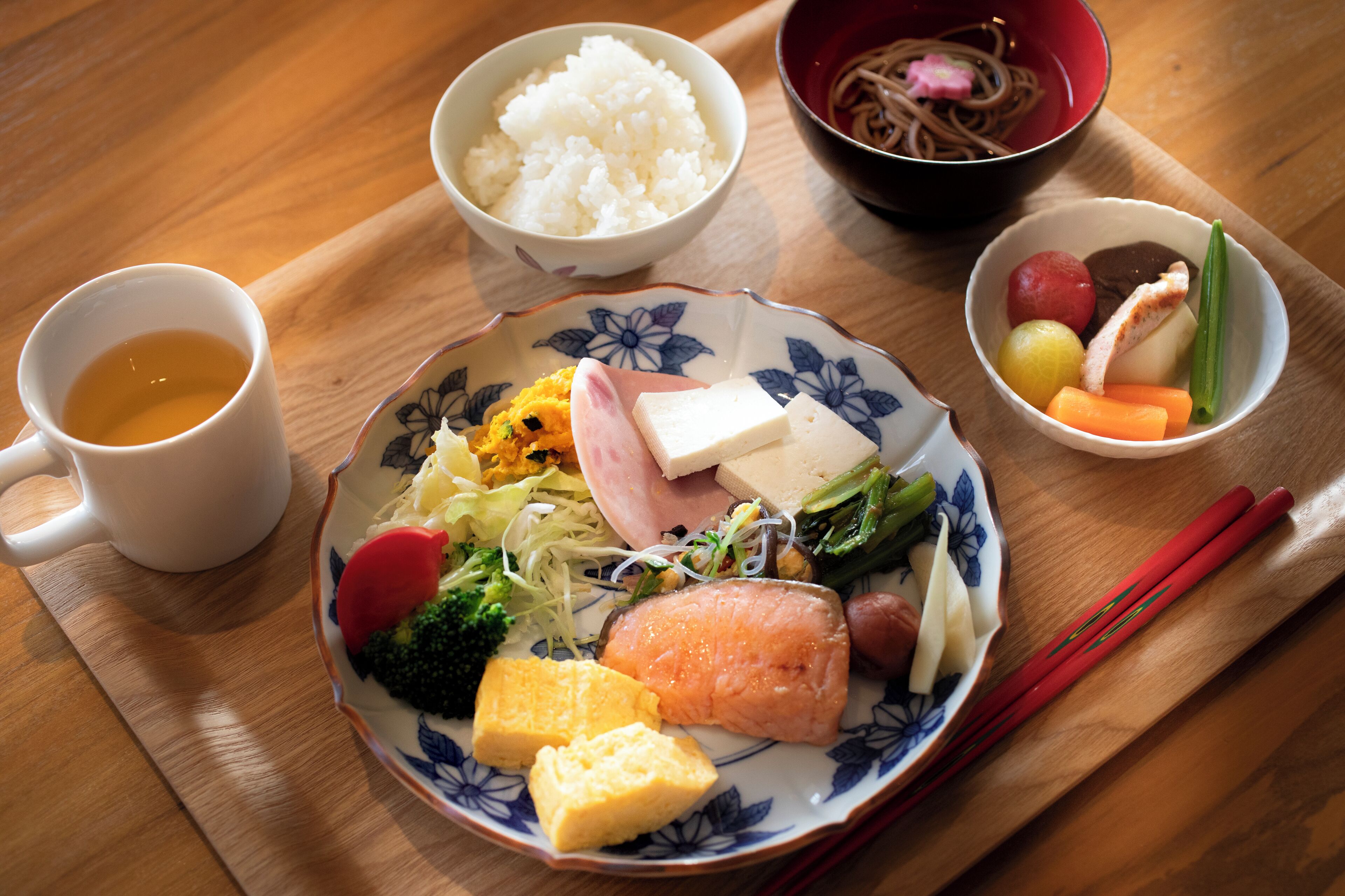 A wooden table with a plate of food, a bowl of rice, a bowl of noodles, and a cup of tea.