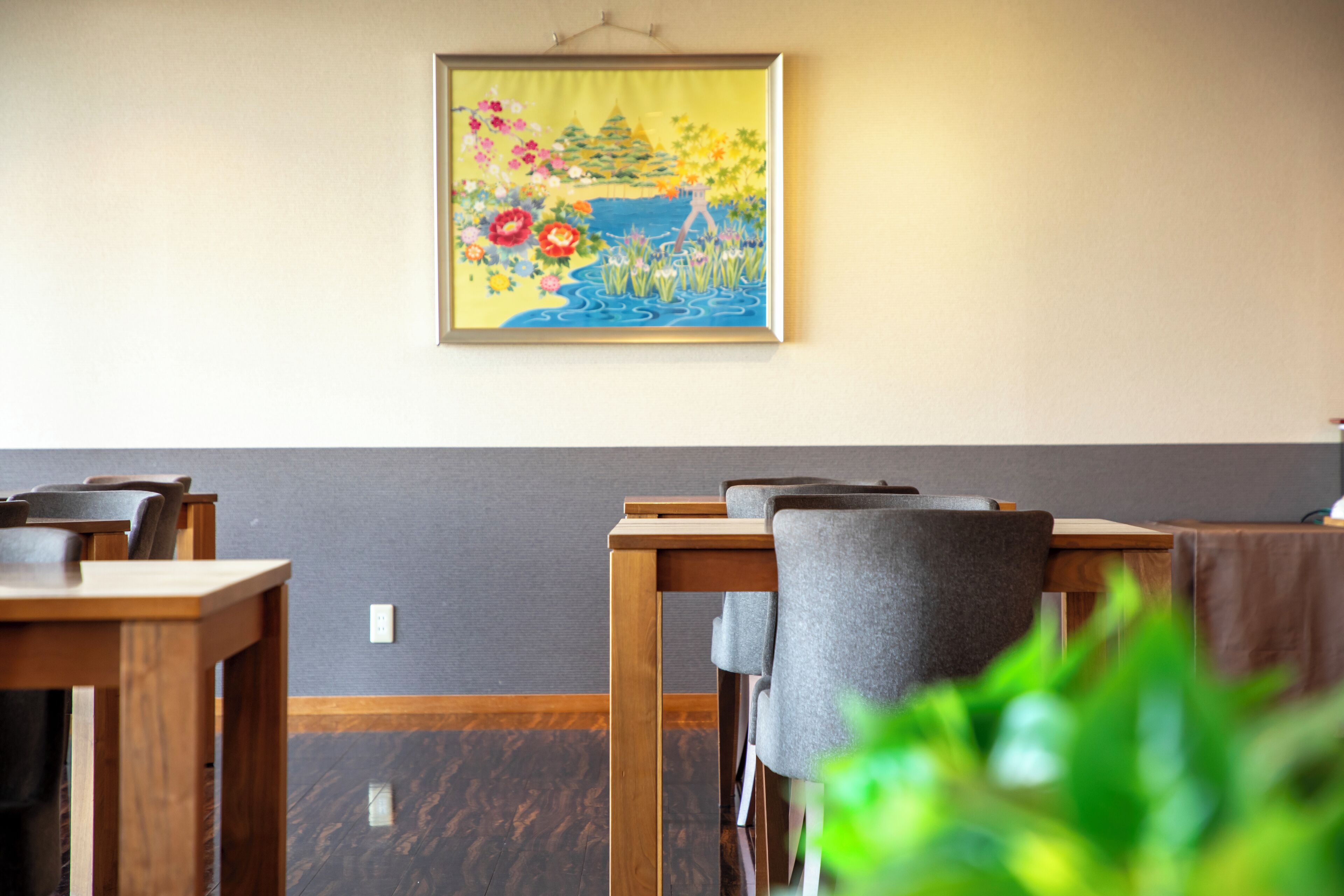 A dining area with wooden tables and chairs, a painting on the wall, and a plant in the foreground.